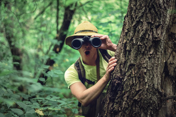 Amazed traveler in hat is looking through binoculars over green forest background.