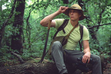 Naklejka na ściany i meble Tired traveler in hat with machetes is sitting on a fallen tree in a green forest.