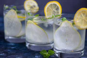 Pear tincture in a transparent glass. Ice cubes and pieces of fruit are also in the middle of the dishes. All on a dark background. Slices of lemon and mint leaves serve as a decoration for the drink.