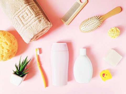 Yellow sponge, beige towel, toothbrush, shampoo, shower gel, wooden hair brushes, soap bar and rubber duck on a pink table. Flat lay photo
