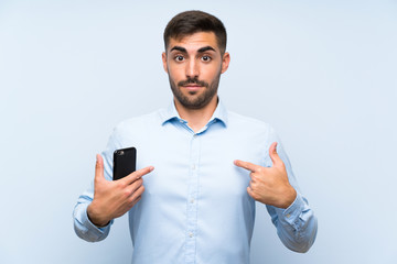 Young handsome man with his mobile over isolated blue wall with surprise facial expression