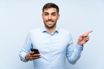 Young handsome man with his mobile over isolated blue wall surprised and pointing finger to the side