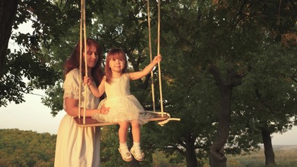 Mom shakes her daughter on swing under tree in sun. close-up. child laughs and rejoices. mother and baby ride on rope swing on an oak branch in forest. Family fun in park, in nature. warm summer day