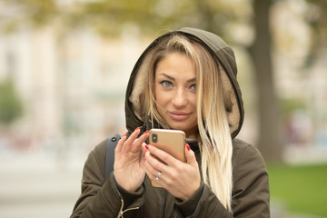 Young beautiful woman using her mobile phone at dusk in the city. Smiling girl sending message from cellphone in urban environment. Woman surfing the net with smartphone on autumn day in the street.