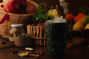 ocktail in a tiki glass on a wooden background. Background decorated with fruits and herbs.