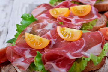 Close-up of thin slices of Italian cold cut pork Coppa on slices of grain bread with salad leaves and cherry tomatoes