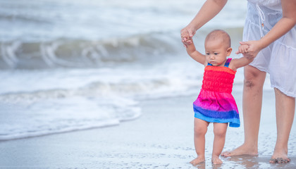 Ten months old baby girl walking on the beach in beautiful summer day. Mother help little baby walking on the beach first time...