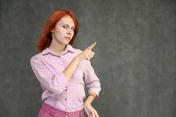 Photo Portrait of a cute female girl manager with bright red hair manager in a pink shirt on a gray background in the studio. He talks, shows his hands in front of the camera with emotions.