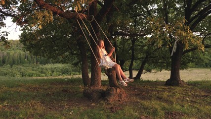 young girl swinging on a swing under a tree in sun, playing with children. Family fun in nature. child rides rope swing on an oak branch in the park the sunset. girl laughs, rejoices.