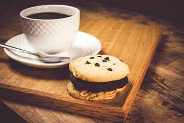 Cookie butter and chocolate Cookies stacked 3 pieces, black coffee in the cup white on white coasters,and stainless steel spoon are on a wooden tray ,  all placed on a wooden table.