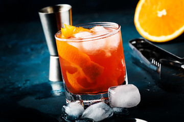 Garibaldi alcoholic cocktail with red bitter, orange juice, zest and ice. Dark blue background, bar tools, selective focus