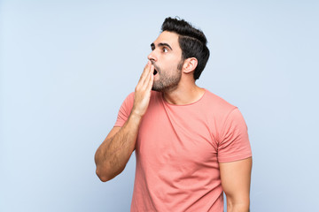 Handsome young man in pink shirt over isolated blue background yawning and covering wide open mouth with hand
