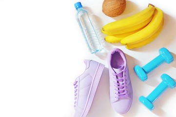 Flat lay fitness items, healthy lifestyle concept. Gym, fitness accessories and proper nutrition. Water bottle, purple sneakers, coconut, a bunch of bananas and blue dumbbells on a white background
