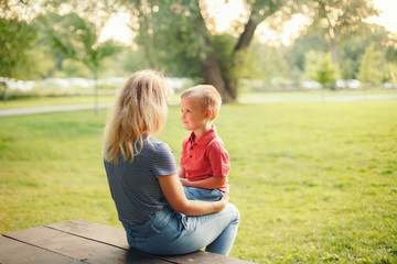 Young blonde Caucasian mother and boy toddler son sitting together face to face on wooden bench outdoor on summer day and talking to each other. Happy family childhood lifestyle.