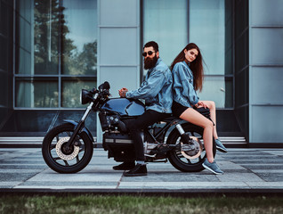 Obraz na płótnie Canvas Beautiful interesting couple is sitting onthe bike in denim jacket near glass building.