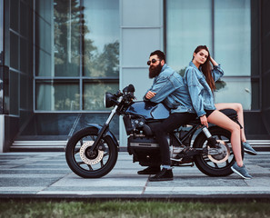 Plakat Beautiful interesting couple is sitting onthe bike in denim jacket near glass building.