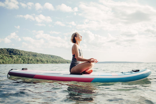 Middle Age Caucasian Woman Practising Yoga On Paddle Sup Surfboard At Sunset. Female Stretching Doing Workout On Lake Water. Modern Individual Hipster Outdoor Summer Sport Activity.