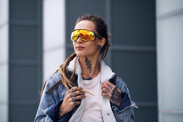 Styilish punk girl wirh dreadlocks, tatooes on her hands and big toned glasses is standing over...