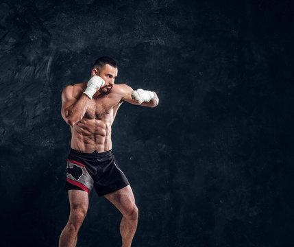 Strong muscular fighter is showing his punch while posing for photographer at dark photo studio.