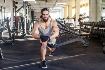 Fototapeta na wymiar Portrait of young adult sportman athlete with long curly hair working out in gym, squating on one knee, stretching after training, doing exercises for legs, squatting. looking at camera and smiling