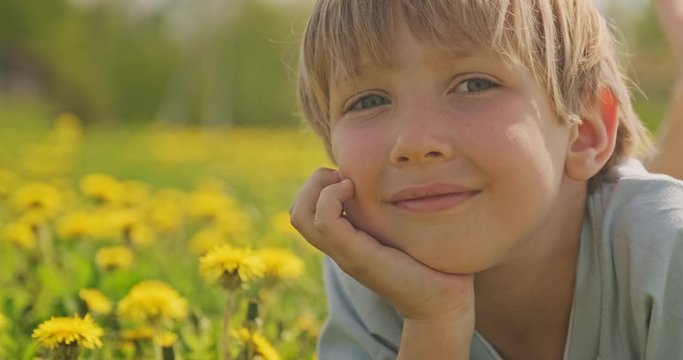 Pretty caucasian boy lying on a spring meadow and looks. Happy child lies on daisies field, closeup view. Cute smiling  face of a blond hair boy, on the meadow. Happiness and childhood. Slow motion