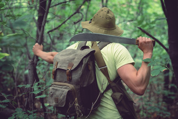 Tourist in hat with a backpack and machetes is walking in forest thickets.