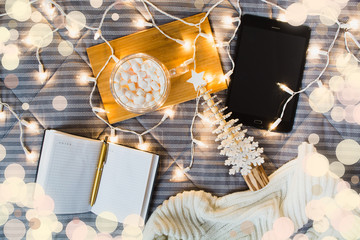 Christmas list of gifts and online shopping concept from home. Flat lay. Wish list or planning with tablet, note, wooden fir tree, marshmallows in mug, lights and cozy home