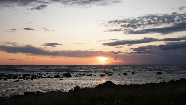 Time lapse showing a beautiful sunset over the ocean with waves hitting the shore while reeds are blowing in the wind.