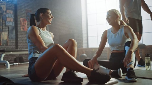 Two Beautiful Fit Athletic Girls Sit on a Floor of Industrial Loft Gym. They're Happy with their Training Program and Successfully Give a High Five. Strong Masculine Man Walks in the Background.