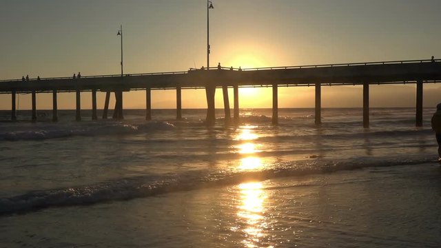 Silhouetted couple walking along the beach by a pier at sunset