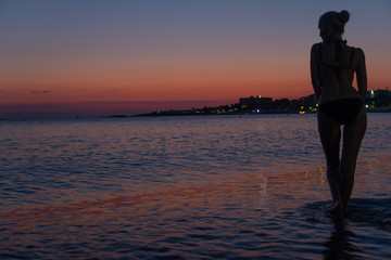 Fototapeta premium Sexy beautiful woman stands on the beach at orange sunset in the warm sea with silhouette
