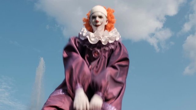A cosplayer with makeup and in a costume like a dancing clown Pennywise dances against the sky with clouds. Cosplay IT next to the fountain. Halloween celebration or All Saints Day masquerade.