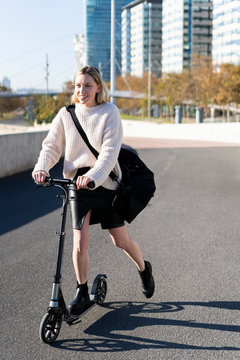 Smiling young woman with sports bag riding kick scooter