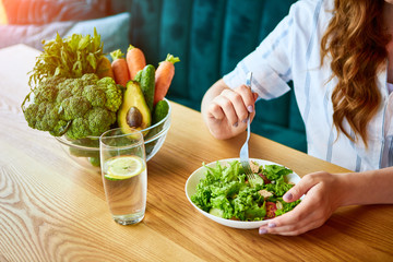 Young happy woman eating salad in the beautiful interior with green flowers on the background and fresh ingredients on the table. Healthy food concept