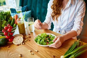 Young happy woman eating salad in the beautiful interior with green flowers on the background and fresh ingredients on the table. Healthy food concept - obrazy, fototapety, plakaty