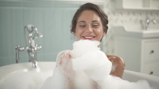 Portrait shot of a young cheerful woman enjoying bubble bath. Girl is smiling and blowing fluffy foam into camera. Spa and wellness concept.