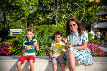 Fototapeta na wymiar Tasty summer obsession concept. Happy young family boys with mother wearing colorful clothes, eating mini melts ice cream in heat cap over summer city park green nature plants background
