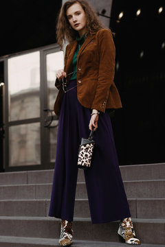 Outdoor full-length street fashion portrait of young curly woman wearing  stylish autumn look: brown corduroy blazer, purple culottes, cowboy boots  with mixed animal prints, holding trendy box bag Stock Photo | Adobe Stock