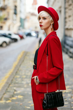 Outdoor autumn fashion portrait of young elegant lady with short blonde hair, red lips makeup, wearing red suit, hat, with small shoulder bag. Model  walking in street of European city