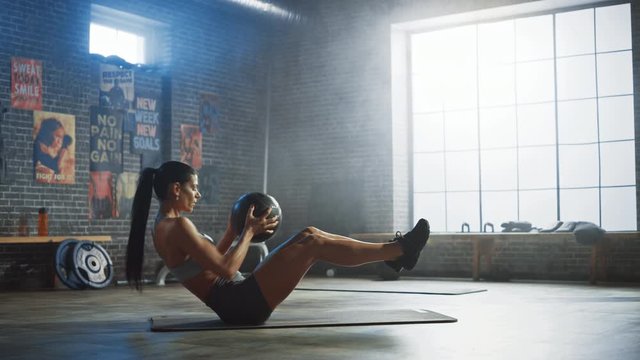 Strong and Fit Beautiful Athletic Woman in Sport Top is Doing Core and Ab Exercises with Ball in a Loft Style Industrial Gym with Motivational Posters. It's Part of Her Cross Fitness Training Workout.