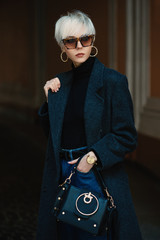  Outdoor fashion portrait of lady with short blonde hair wearing classic gray coat, black...