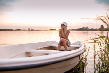 Young woman in a dress sitting on the boat and enjoying in a beautiful sunset while drinking beer