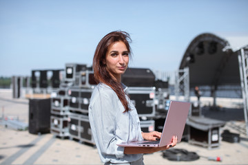 Installation of stage equipment and preparing for a live concert open air. Event manager portrait. Summer music city festival. Young serious woman stand and work with her laptop near the stage.