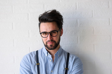Portrait of handsome bearded hipster guy with glasses on standing indoors by the white brick wall