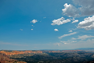 Look over valley at Bryce Canyon with mountains full of trees and clouds in the blue sky