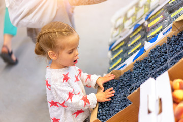 Little Caucasian beautiful girl is very surprised when choosing berries in a large store. Cute child is looking at blueberries.
