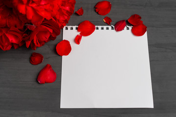 Empty sheet of paper for text next to a bouquet and red rose petals against a dark background.
