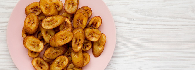 Homemade fried plantains on a pink plate on a white wooden surface, top view. Flat lay, overhead, from above. Space for text.