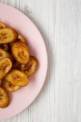 Homemade fried plantains on a pink plate on a white wooden surface. Flat lay, overhead, from above. Copy space.