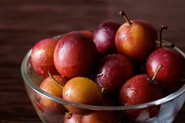 Ripe sweet plum fruits with water drops in glass bowl on dark moody wood table background
